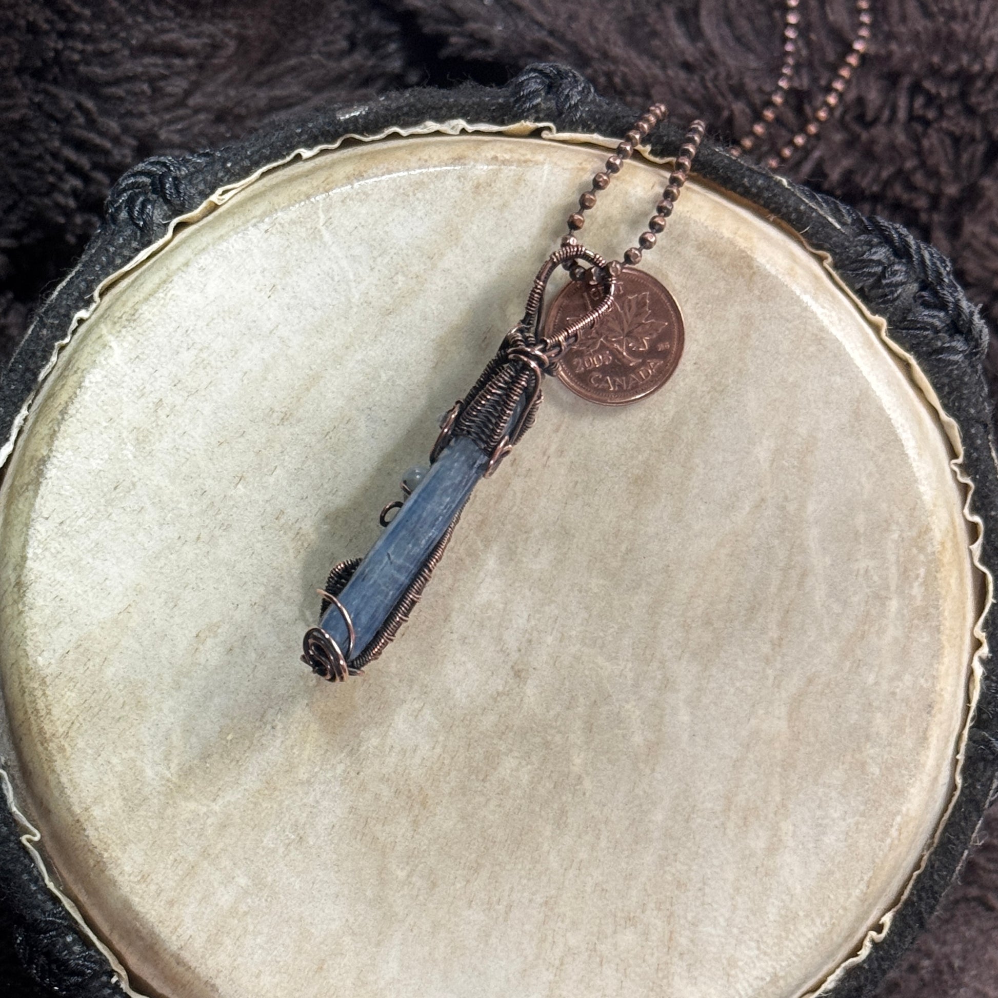 Narrow side profile of a polished blue kyanite blade showing its natural striations, slim depth, patinated copper frame and woven bail.