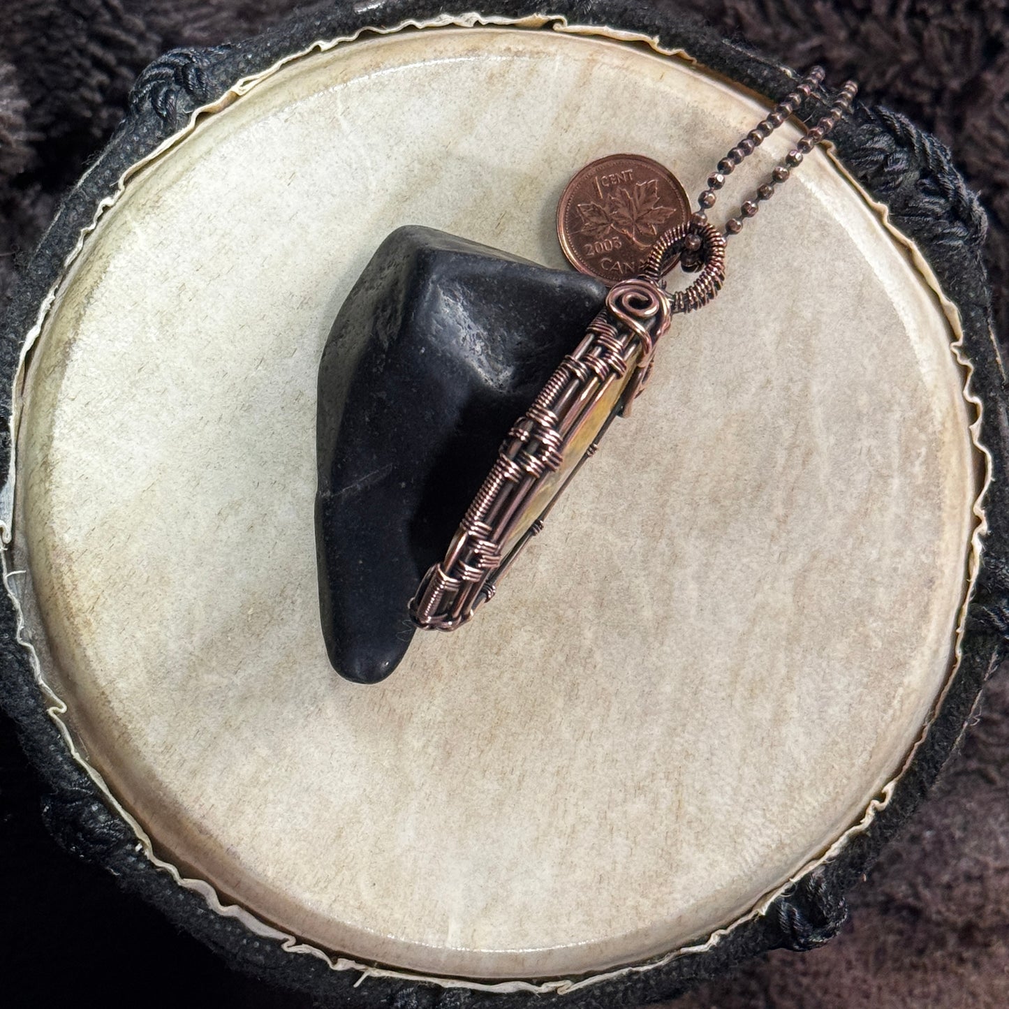 Side profile of a Bumble Bee Jasper pendant showing the depth of the cabochon, coiled copper frame and woven bail, styled against shungite.