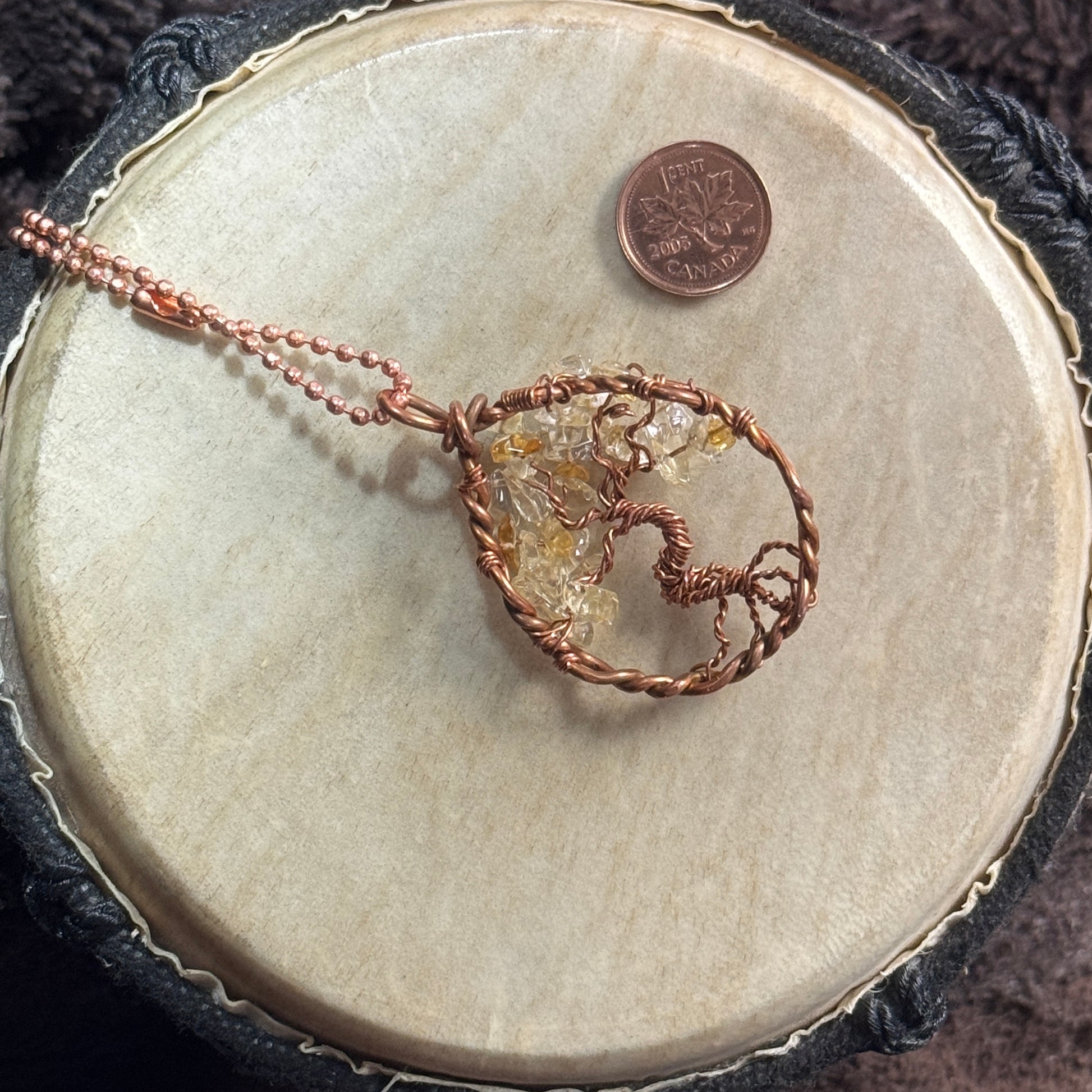 Reverse view of a citrine Tree of Life pendant showing its windswept copper trunk, exposed roots and teardrop frame beside a Canadian penny.