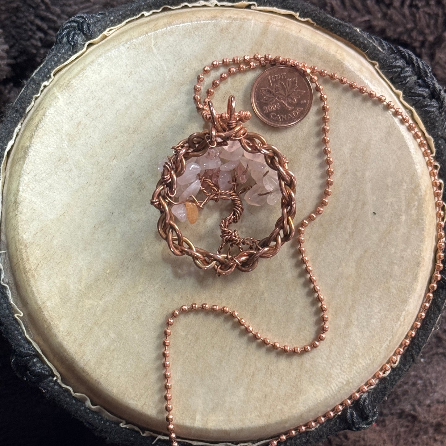 Reverse view of a petite rose quartz Tree of Life pendant showing its twisted copper trunk, woven frame and faceted ball chain beside a penny.