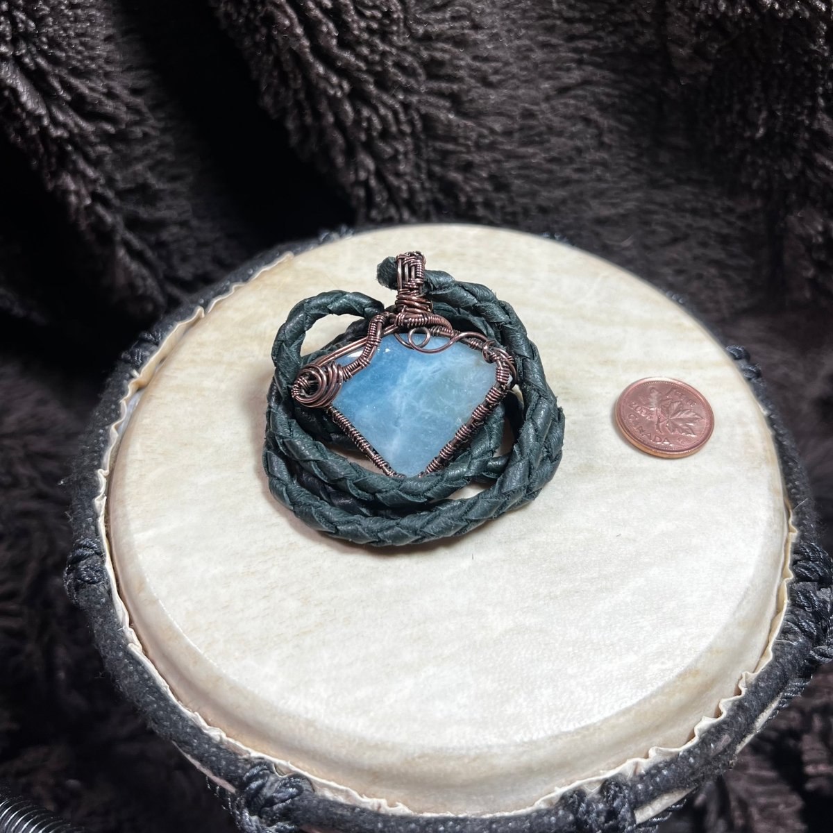 Blue stone pendant with braided design on a round white stone surface, next to a penny for scale.
