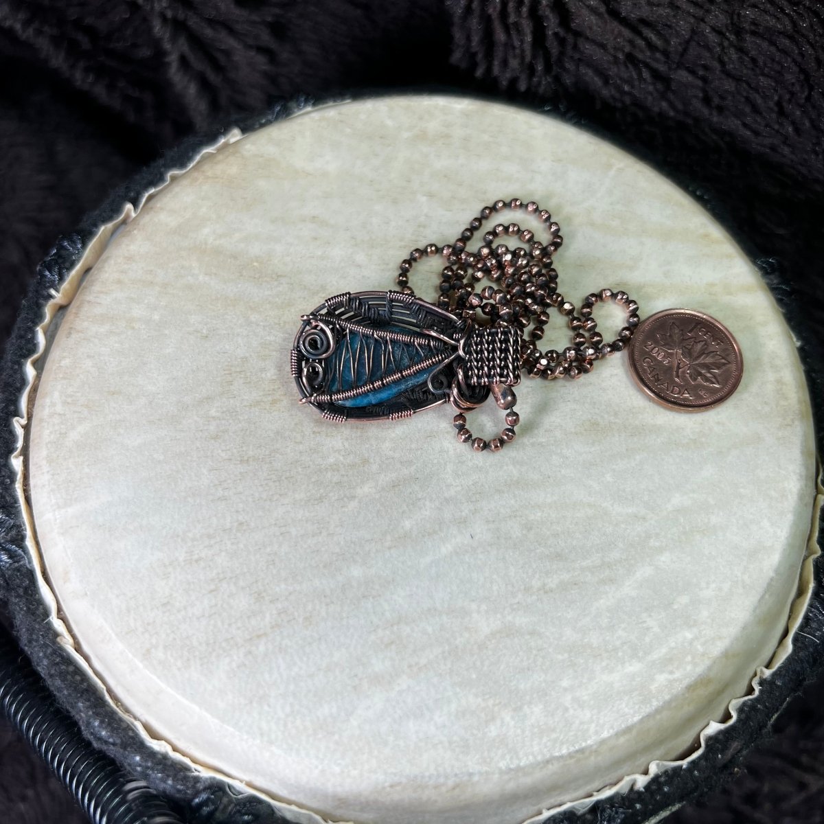Back side of a necklace with a blue pendant on a round white surface next to a penny for scale