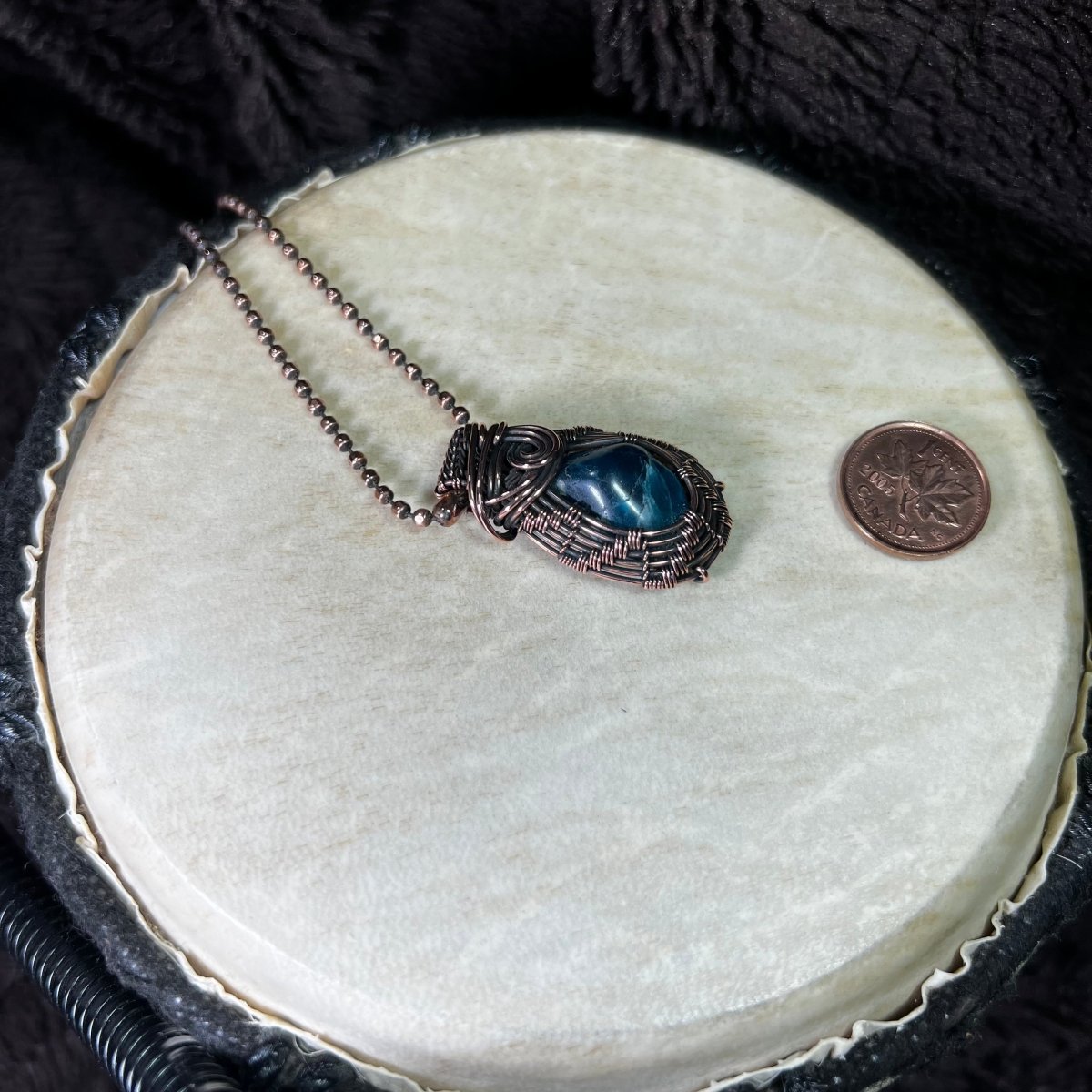 Necklace with a blue stone pendant on a white circular surface next to a penny.
