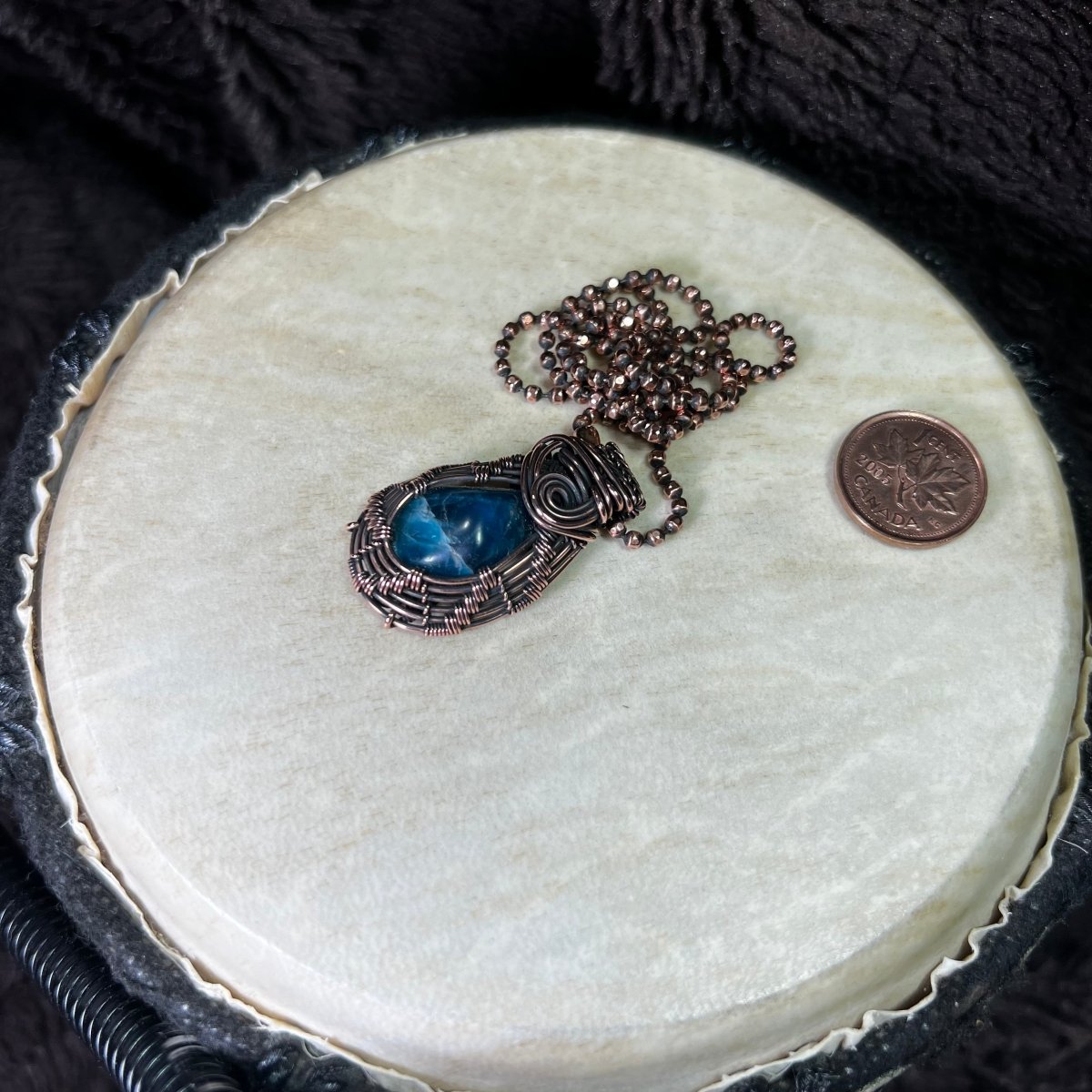 Necklace with a blue stone pendant on a round white surface, next to a penny for scale.