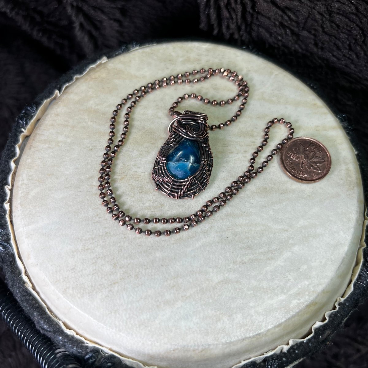 Necklace with a blue pendant on a round white surface next to a penny for scale