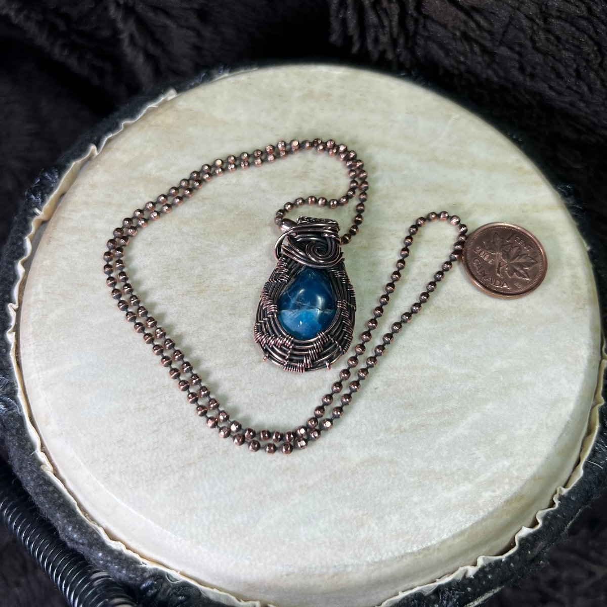 Necklace with a blue pendant on a round white surface next to a penny for scale