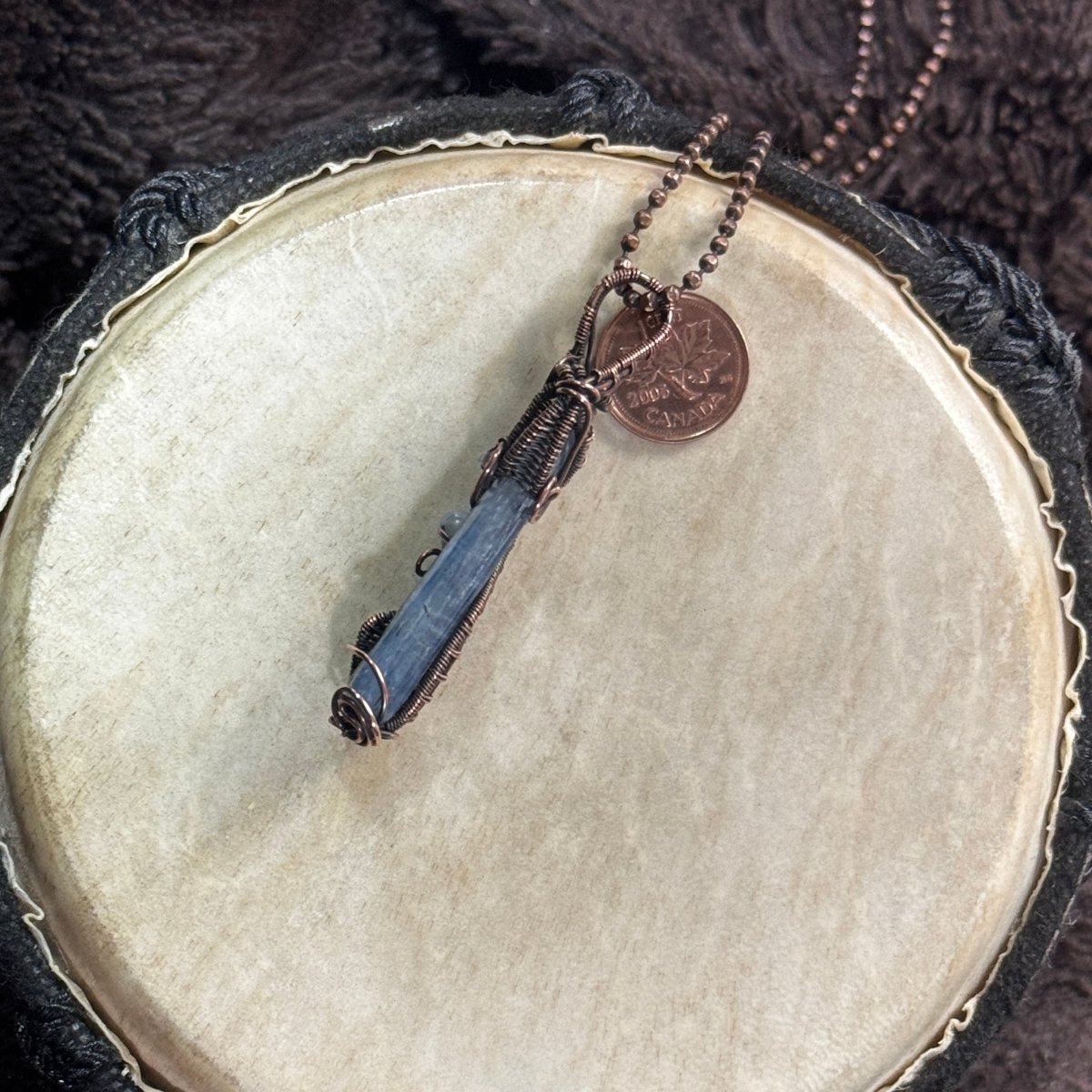 Narrow side profile of a polished blue kyanite blade showing its natural striations, slim depth, patinated copper frame and woven bail.