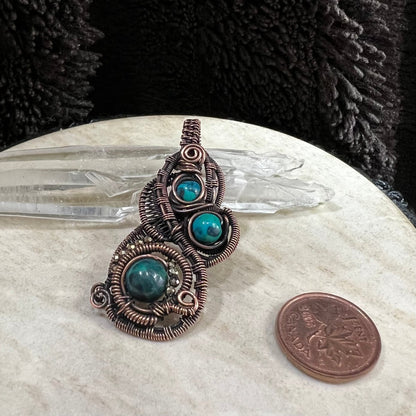 Antiqued copper wirework pendant with chrysocolla beads on a white surface next to a Canadian penny for scale.