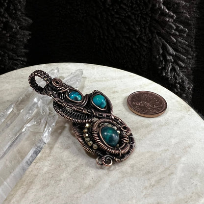 Antiqued copper intricate wirework pendant with chrysocolla beads on a white surface next to a Canadian penny for scale.