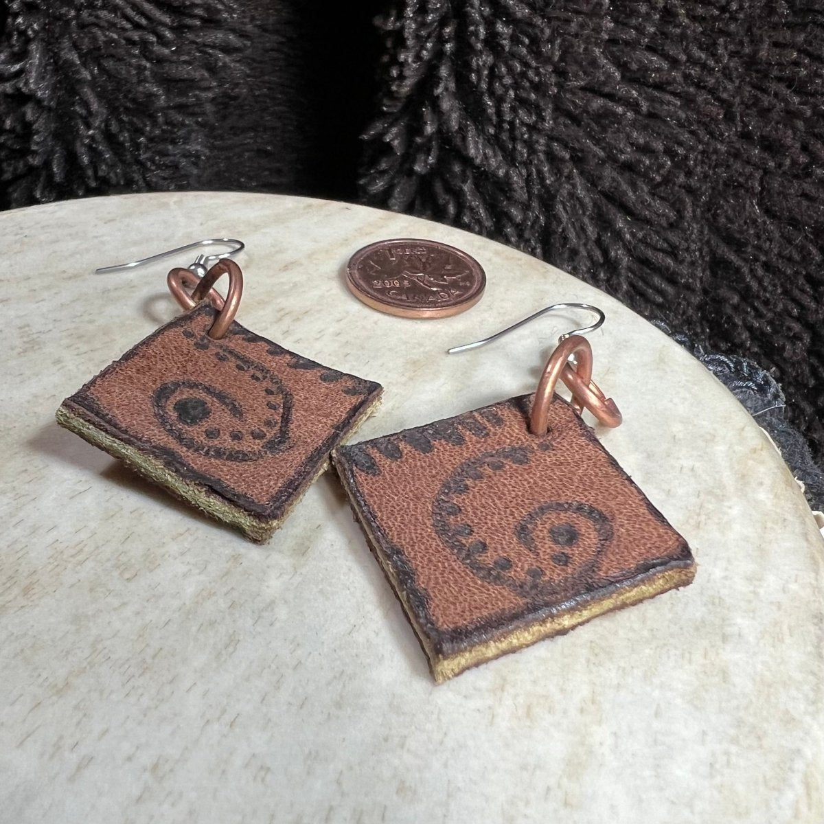 Handmade earrings crafted from reclaimed leather belt pieces with hand-burned spiral designs, copper rings, and surgical steel ear wires, photographed with Canadian penny for scale.