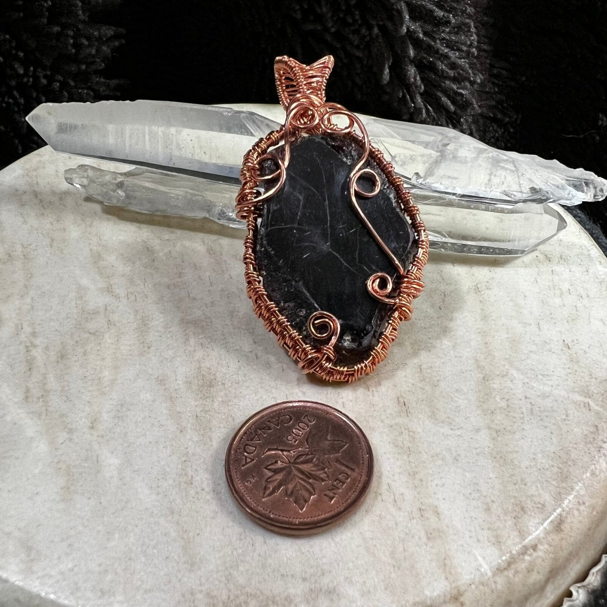 Garnet slice pendant with copper wire wrapping on a beige surface with crystals in the background next to a Canadian pennt for scale