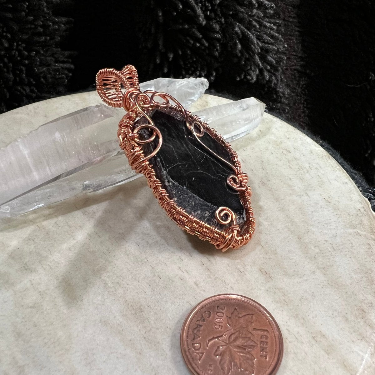 Garnet slice pendant with copper wire wrapping on a beige surface with crystals in the background next to a Canadian pennt for scale