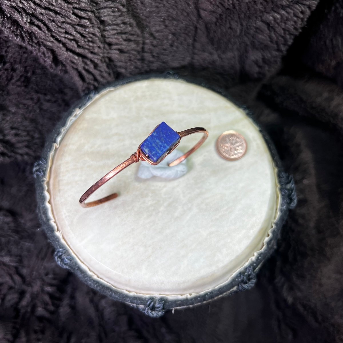 Copper bracelet with a blue lapis lazuli stone on a round white surface, next to a penny for scale.