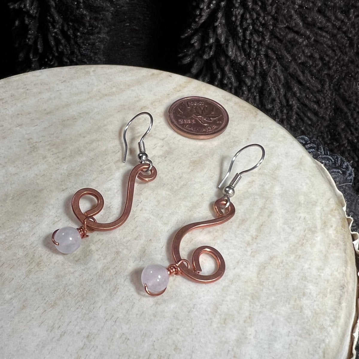Handmade hammered copper spiral earrings with rose quartz bead drops and surgical steel ear wires, photographed with Canadian penny for scale.