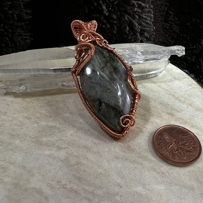 Labradorite pendant with  copper wire wrapping on a neutral surface with a CAnadian penny for scale.