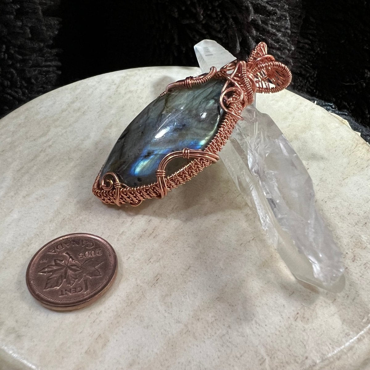 Copper-wrapped labradorite stone pendant on a beige surface with a clear crystal and penny for scale.