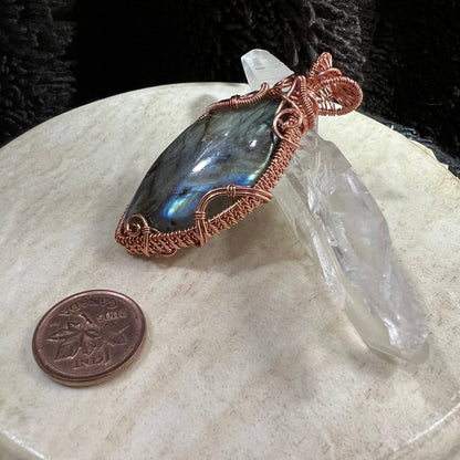Copper-wrapped labradorite stone pendant on a beige surface with a clear crystal and penny for scale.