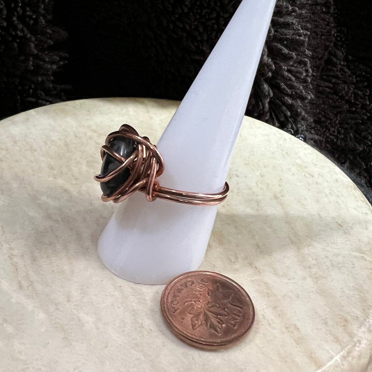 Handmade copper wire-wrapped ring with a black stone on a white candle, next to a penny for scale.