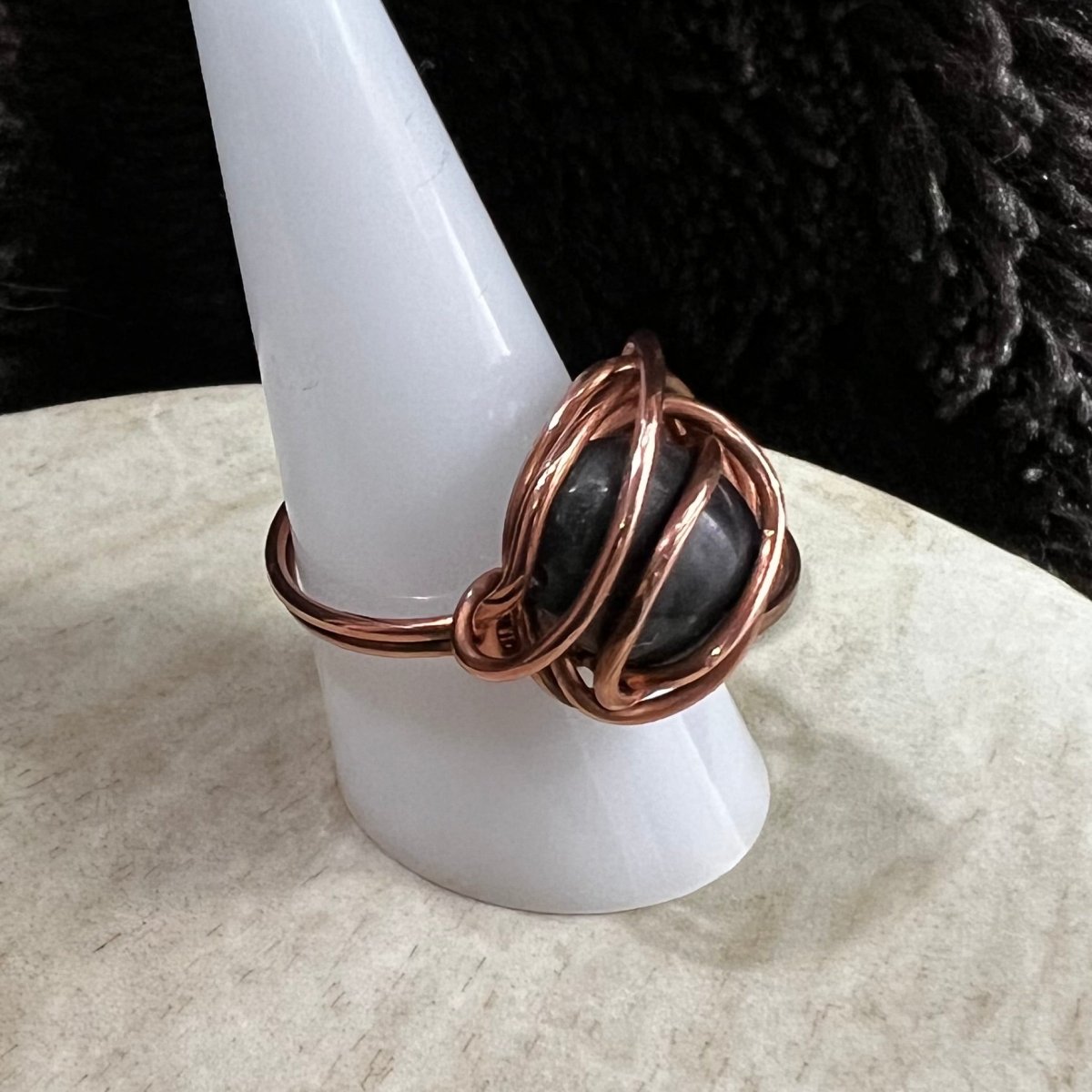 Copper wire-wrapped ring with a central  larvikite stone on a white pedestal against a dark background