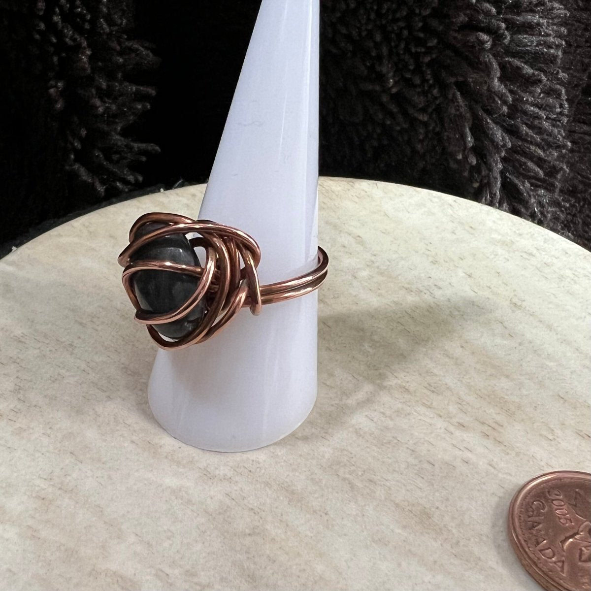 Copper wire-wrapped ring with black larvikite stone on a white stand, next to a penny for scale.