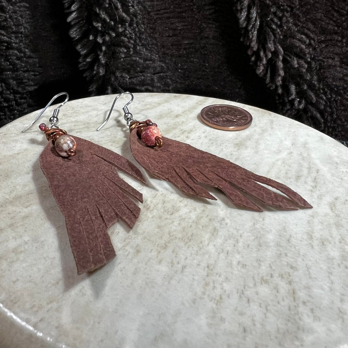 Brown leather tassel earrings on a white surface with a coin for scale.