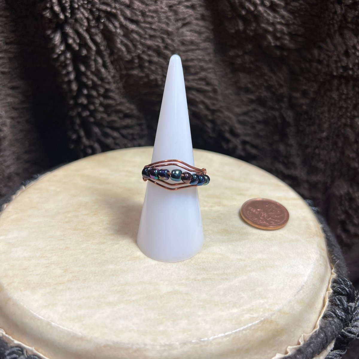 Beaded ring on a white cone-shaped stand with a penny for scale, against a textured brown background.