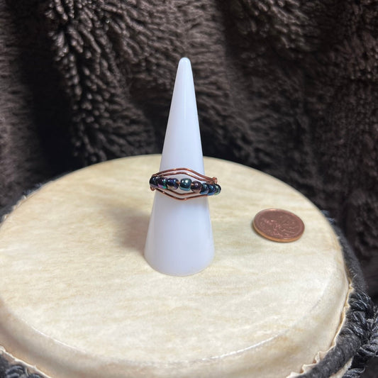 Beaded ring on a white cone-shaped stand with a penny for scale, against a textured brown background.