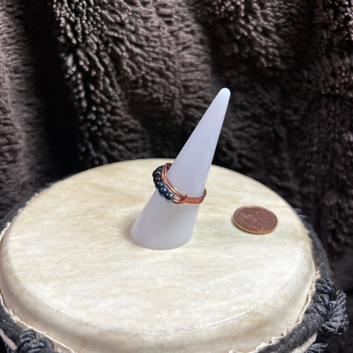 Beaded ring on a white cone-shaped stand with a penny for scale, against a textured brown background.
