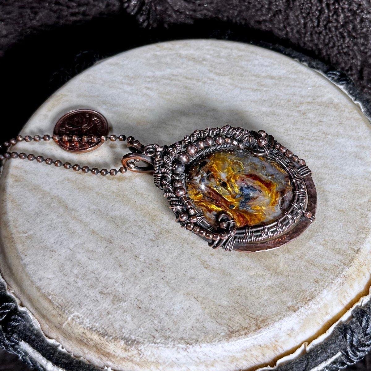 Close front view of a golden and blue Pietersite cabochon surrounded by ornate patinated copper wirework on a faceted copper ball chain.