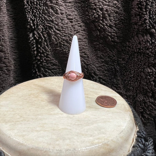 White cone-shaped object with a pink stone ring on top, placed on a round beige surface with a brown textured background.