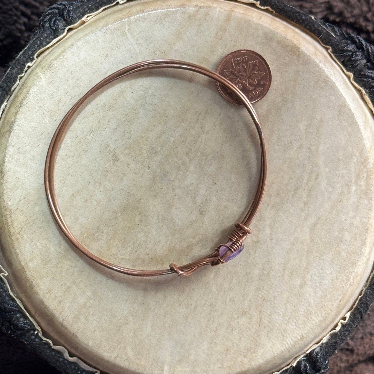 Full view of a three-inch closed reclaimed copper bangle with a small purple amethyst bead, photographed beside a Canadian penny for scale.
