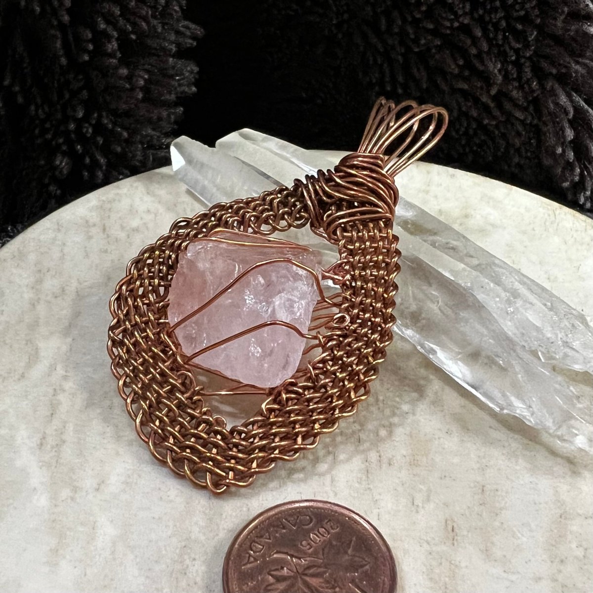 Top view of a raw rose quartz pendant wrapped in handwoven reclaimed copper frame, leaning on 2 quartz points, on neutral background, next to Canadian penny