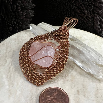 Top view of a raw rose quartz pendant wrapped in handwoven reclaimed copper frame, leaning on 2 quartz points, on neutral background, next to Canadian penny