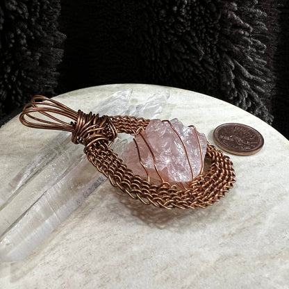 Side view of raw rose quartz pendant wrapped in handwoven reclaimed copper frame, soft pink natural crystal necklace on neutral background