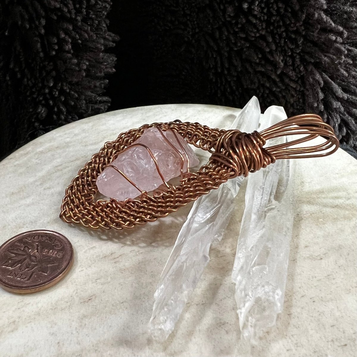 Copper-wrapped raw rose quartz crystal pendant on a neutral surface with a Canadian penny for scale.