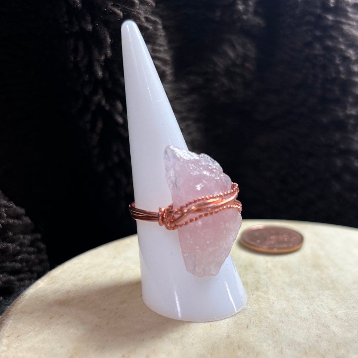 Rose quartz crystal ring with copper wire wrap on a white cone against a dark background