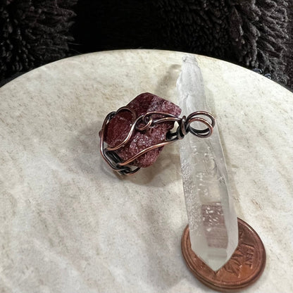 Wire-wrapped red stone on a clear crystal with a penny for scale on a rawhide surface.