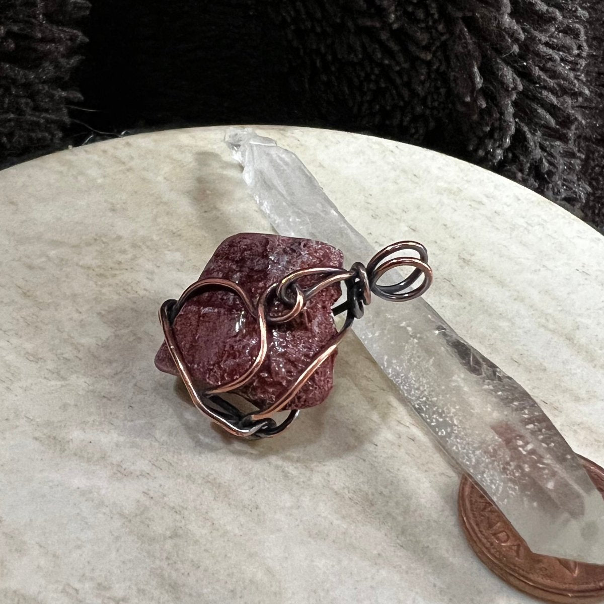 Wire-wrapped raw red calcite stone pendant on a beige surface with a crystal and penny for scale.