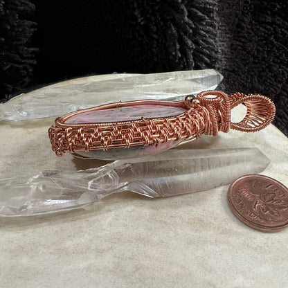 Copper wire-wrapped rhodochrosite pendant leaning on a quartz crystal point next to a Canadian penny for scale