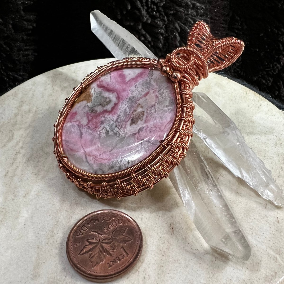 Copper-wrapped  pink rhodochrosite on a beige surface with a clear crystal and Canadian penny for scale.