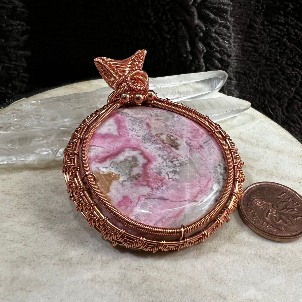 Marble-patterned rhodochrosite palm stone pendant in a copper frame on a marble surface with a Canadian penny  for scale.