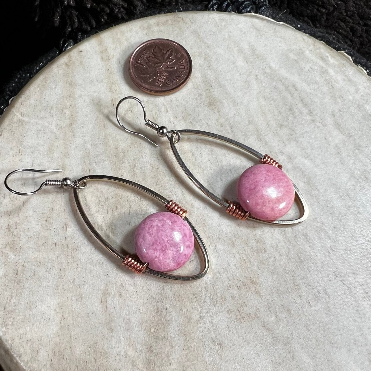 Handmade rhodochrosite bead earrings suspended in reclaimed silver-plated oval hoops with copper wire accents and surgical steel ear wires, photographed with Canadian penny for scale.