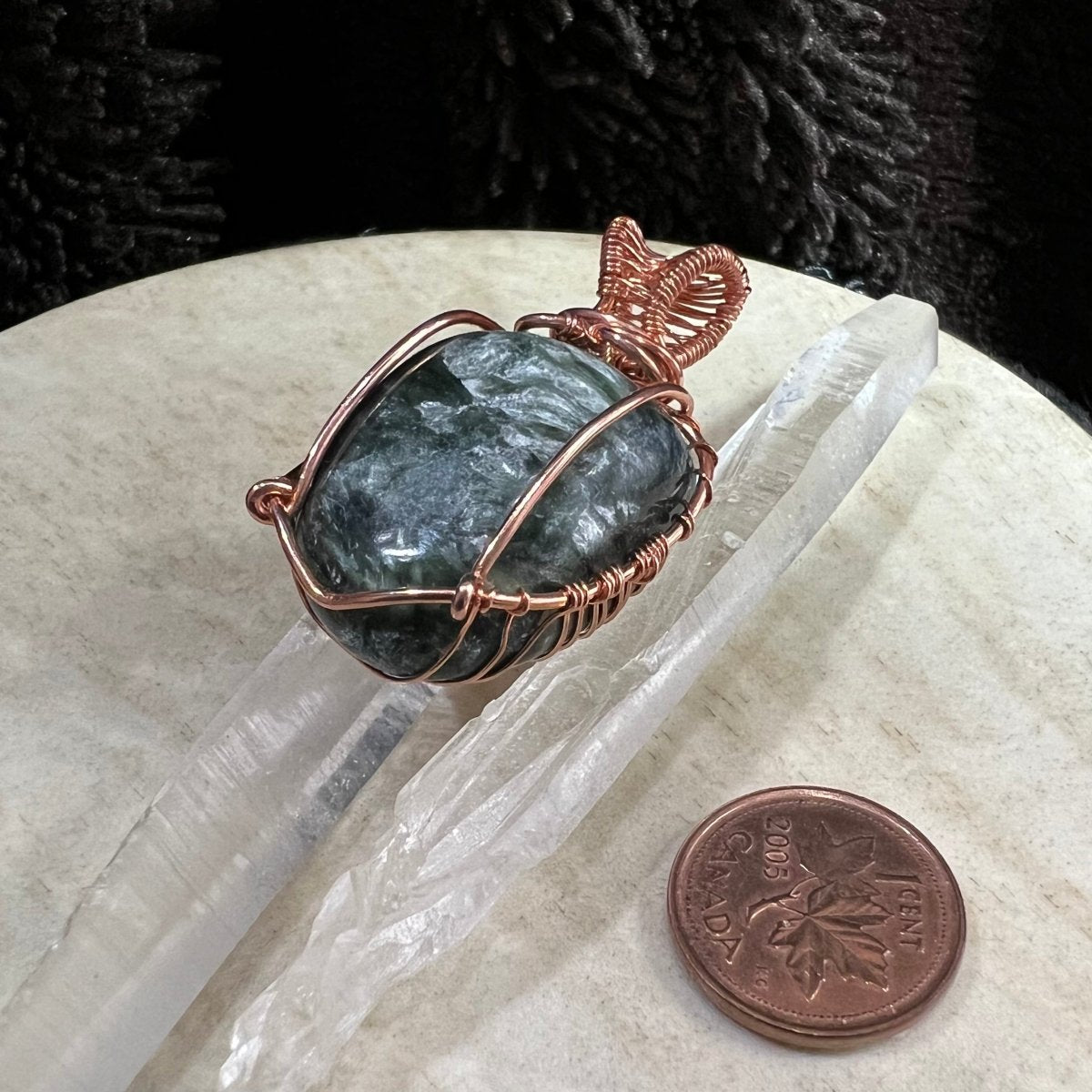Pendant with a seraphinite stone wrapped in copper wire on a neutral surface, next to a penny for scale.