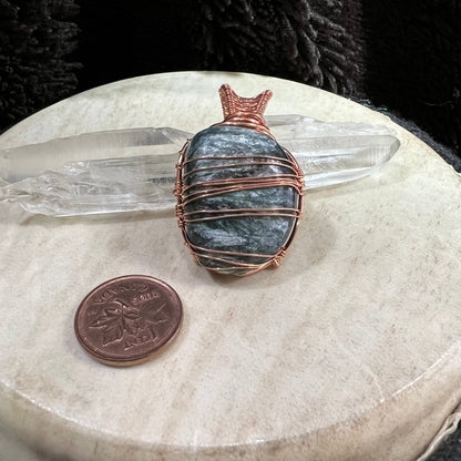 Decorative seraphinite stone with copper wire wrapping on a neutral surface with a crystal and penny for scale.
