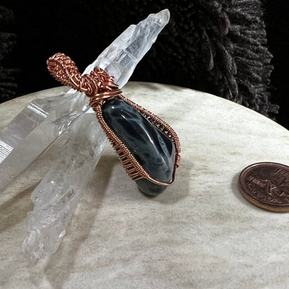 Copper-wrapped spiderweb obsidian crystal on a white surface, next to a Canadian penny for scale. side view.