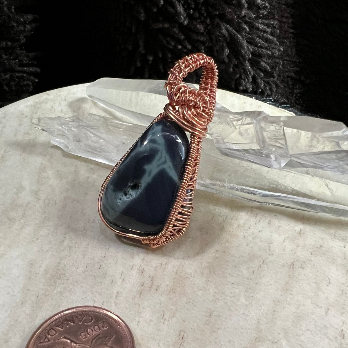 Black stone pendant with copper wire wrapping on a white surface with a penny for scale.