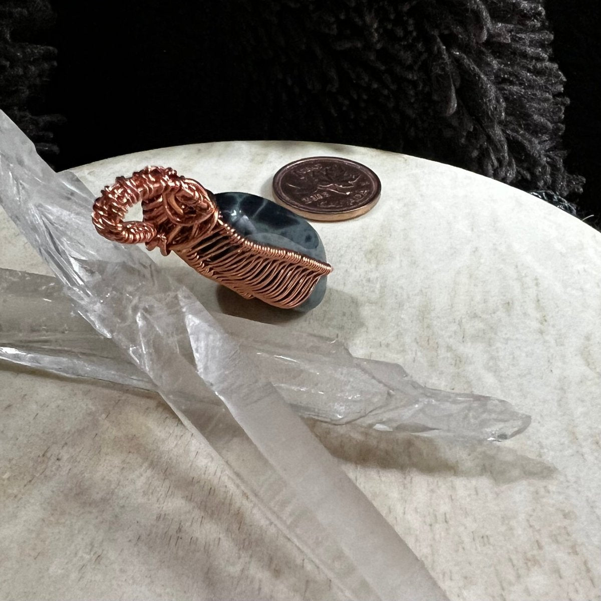 Copper wire wrapped spiderweb obsidian stone on a neutral surface with a Canadian penny for scale.