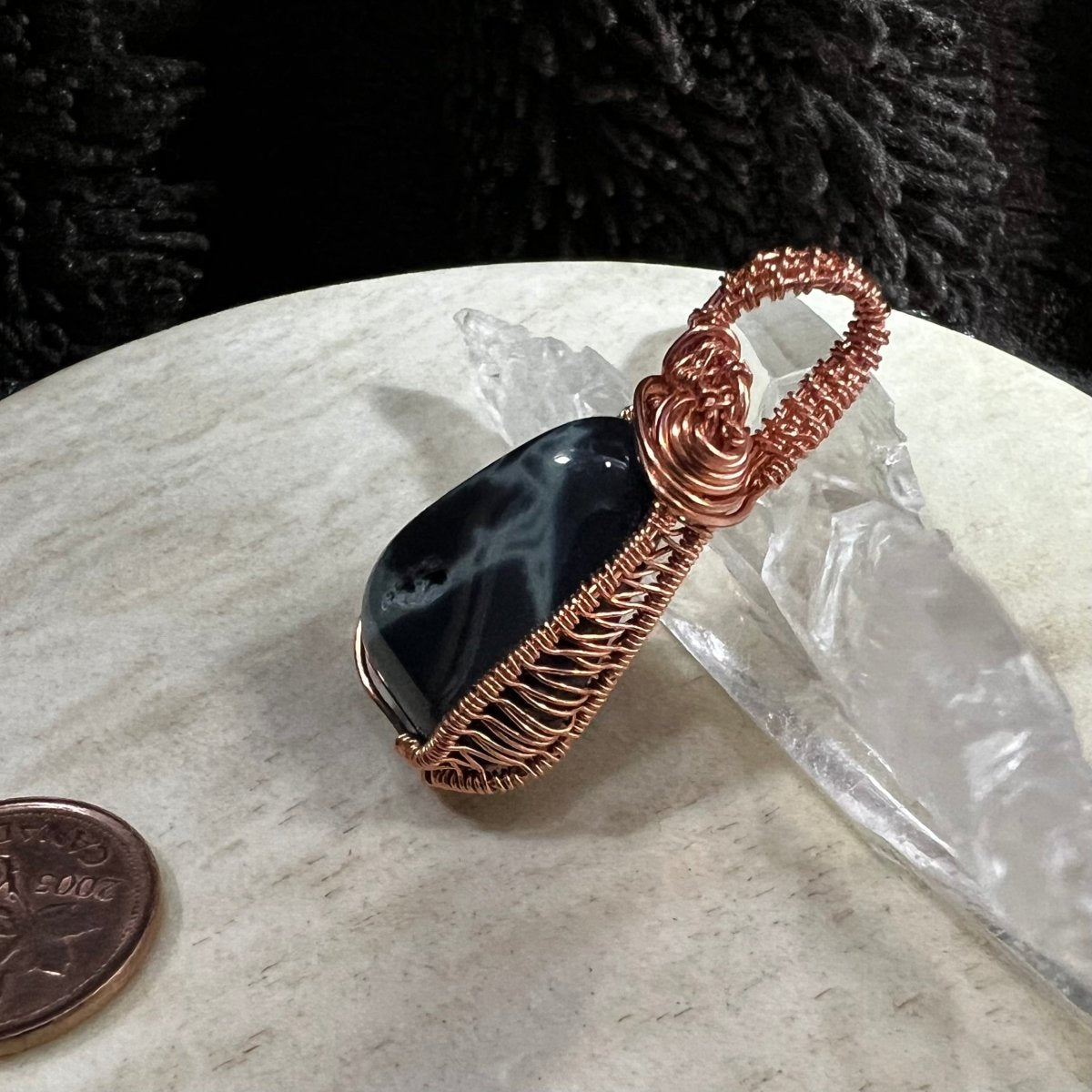 Black and gray stone pendant with copper wire wrapping on a neutral surface next to a Canadian penny.
