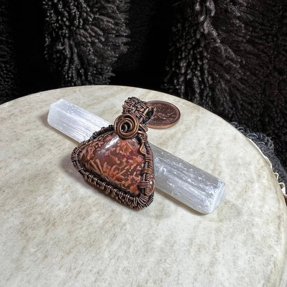 One-of-a-kind Starburst Jasper pendant wrapped in reclaimed copper wire displayed on a selenite wand with penny for size reference.