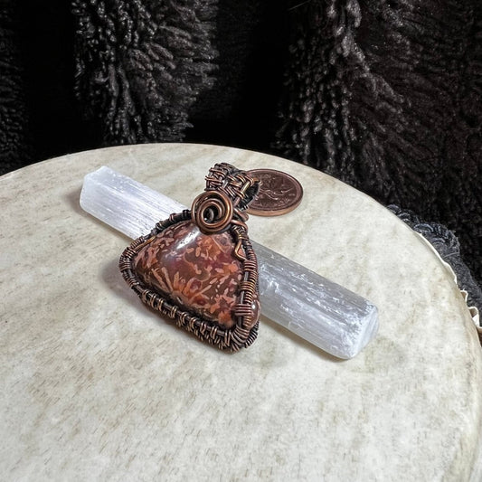 One-of-a-kind Starburst Jasper pendant wrapped in reclaimed copper wire displayed on a selenite wand with penny for size reference.